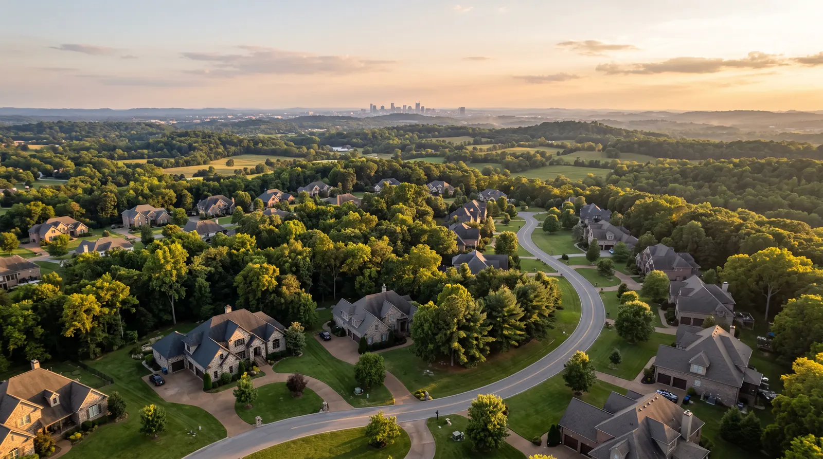 Aerial View of Middle Tennessee Neighborhoods | Prodigy Plumbing & Drain An aerial view over a green Middle Tennessee neighborhood at golden hour showing brick and stone homes, mature trees and rolling hills, a winding road and a glimpse of a city skyline far away
