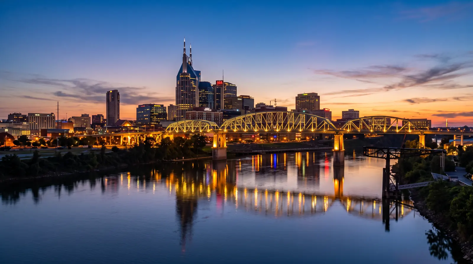 Nashville Skyline at Blue Hour | Prodigy Plumbing & Drain The Nashville skyline glowing at blue-hour dusk beyond the Cumberland River with a calm reflection, a warm sunset gradient in the sky and the lit pedestrian bridge
